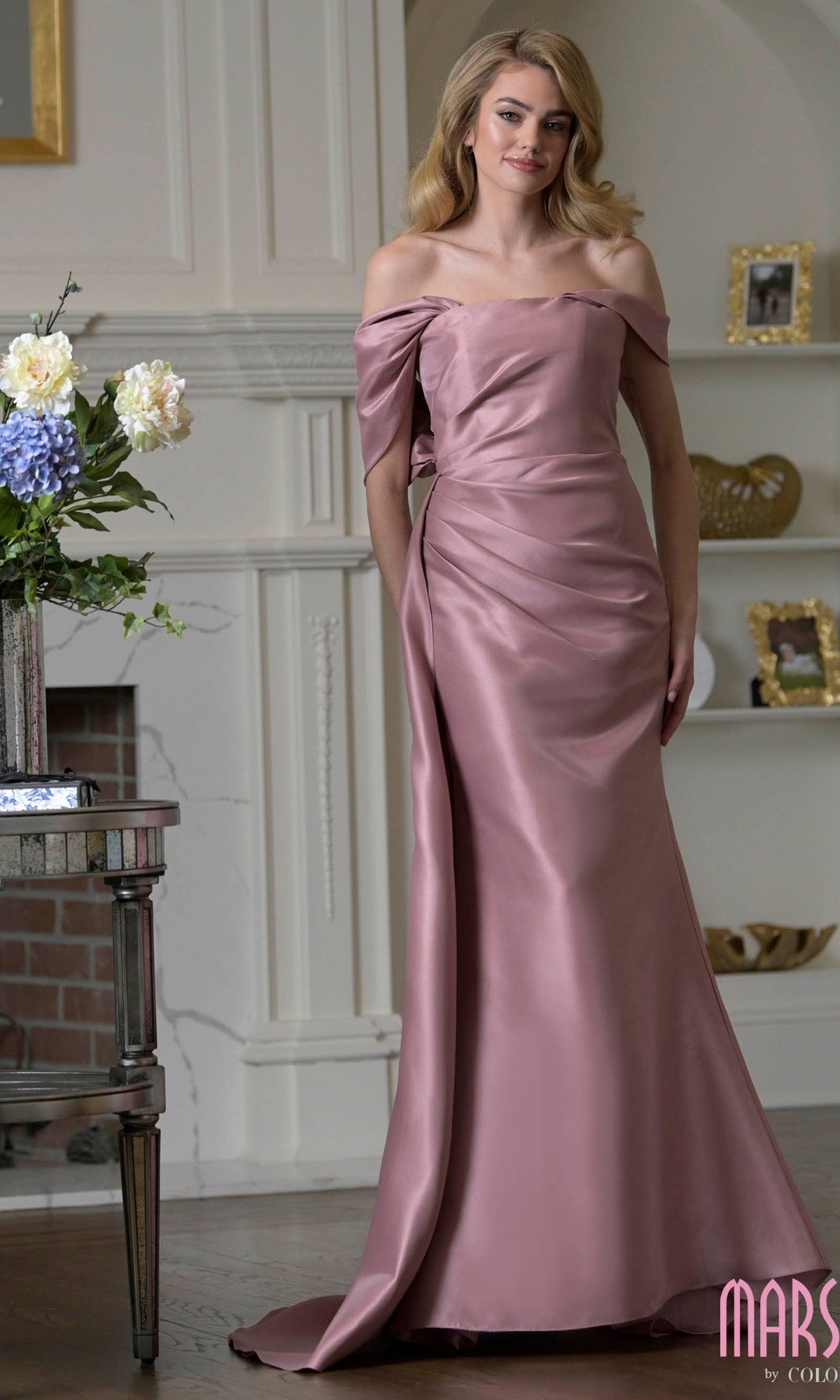 Woman in a Marsoni MV1405 long formal dress in Rose Wood, with an asymmetric neckline and beaded applique. Standing indoors near a fireplace and floral arrangement, her off-the-shoulder, floor-length satin gown radiates elegance.